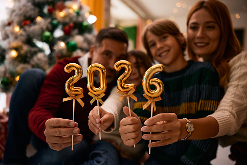 Family celebrating christmas together and holding golden balloons shaped like numbers forming 2026, the new year, sitting in front of a decorated christmas tree